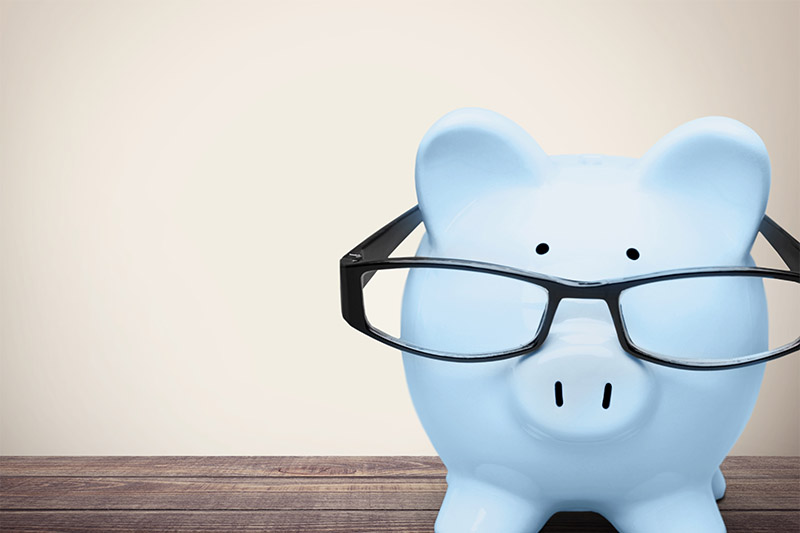 A light blue piggy bank wearing black eyeglasses sits on a wooden surface against a plain, beige background.