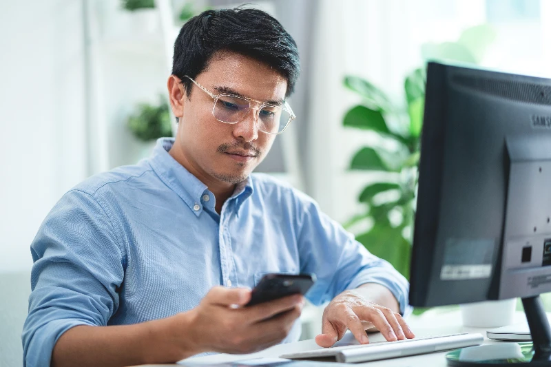 A-man-wearing-glasses-is-sitting-at-a-desk-with-a-computer-monitor-and-a-cell-phone-He-is-looking-at-the-cell-phone-while-typing-on-the-computer
